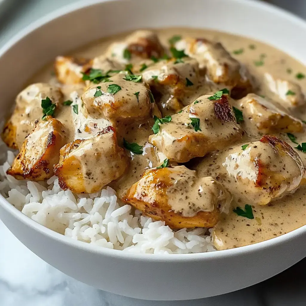 A bowl of rice topped with pieces of chicken coated in a creamy sauce, garnished with fresh parsley.